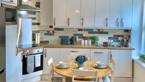 A modern kitchen with white cabinets, a wooden dining table, and a built-in oven. - Stunning 3-bed Apartment in Ventnor (Ventnor)