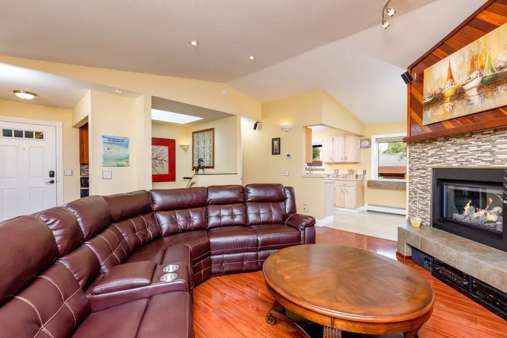 A living room with a brown leather sectional sofa, a round wooden coffee table, and a fireplace with a stone surround.