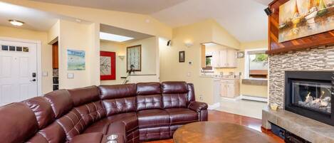 A living room with a brown leather sectional sofa, a round wooden coffee table, and a fireplace with a stone surround.