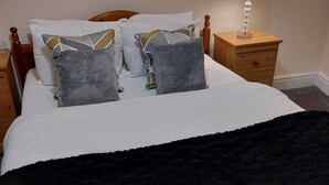 En snyggt bäddad säng med en svart pläd, en sänggavel i trä och flera kuddar. Bredvid sängen står ett litet bord i trä med en lampa på.