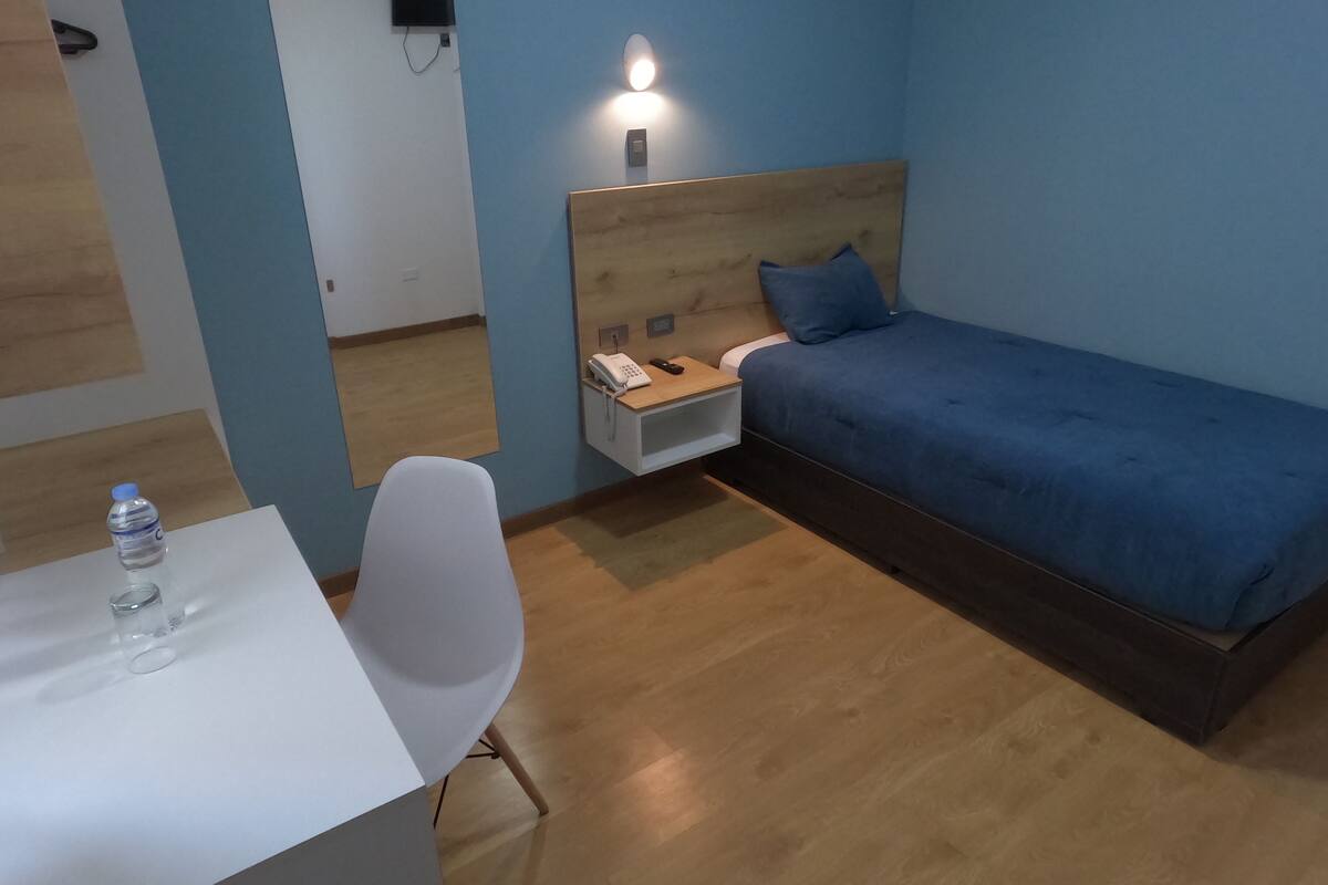 Habitación de hotel con una cama individual, una mesita de noche con teléfono, un escritorio pequeño con silla y una botella de agua sobre el escritorio.