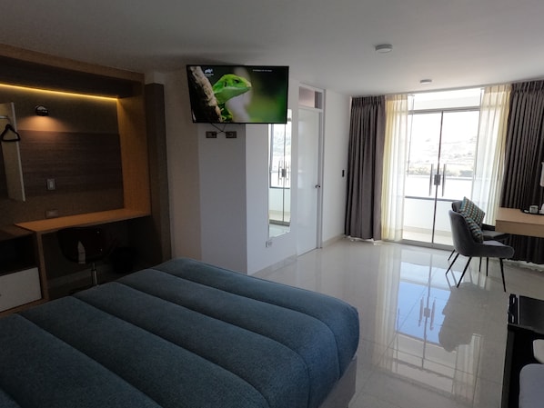 A modern hotel room with a large bed, a flat-screen TV mounted on the wall, a desk with a chair, and a balcony with curtains.