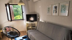 A living room with a grey sofa, a black TV on a white stand, a black chair with a cushion, and a window with a view of greenery. - Apartments Tea Medulin - One Bedroom Apartment with Terrace (Medulin)
