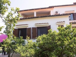 A two-story house with a balcony, a flat roof, and a small garden area with trees and a purple umbrella.