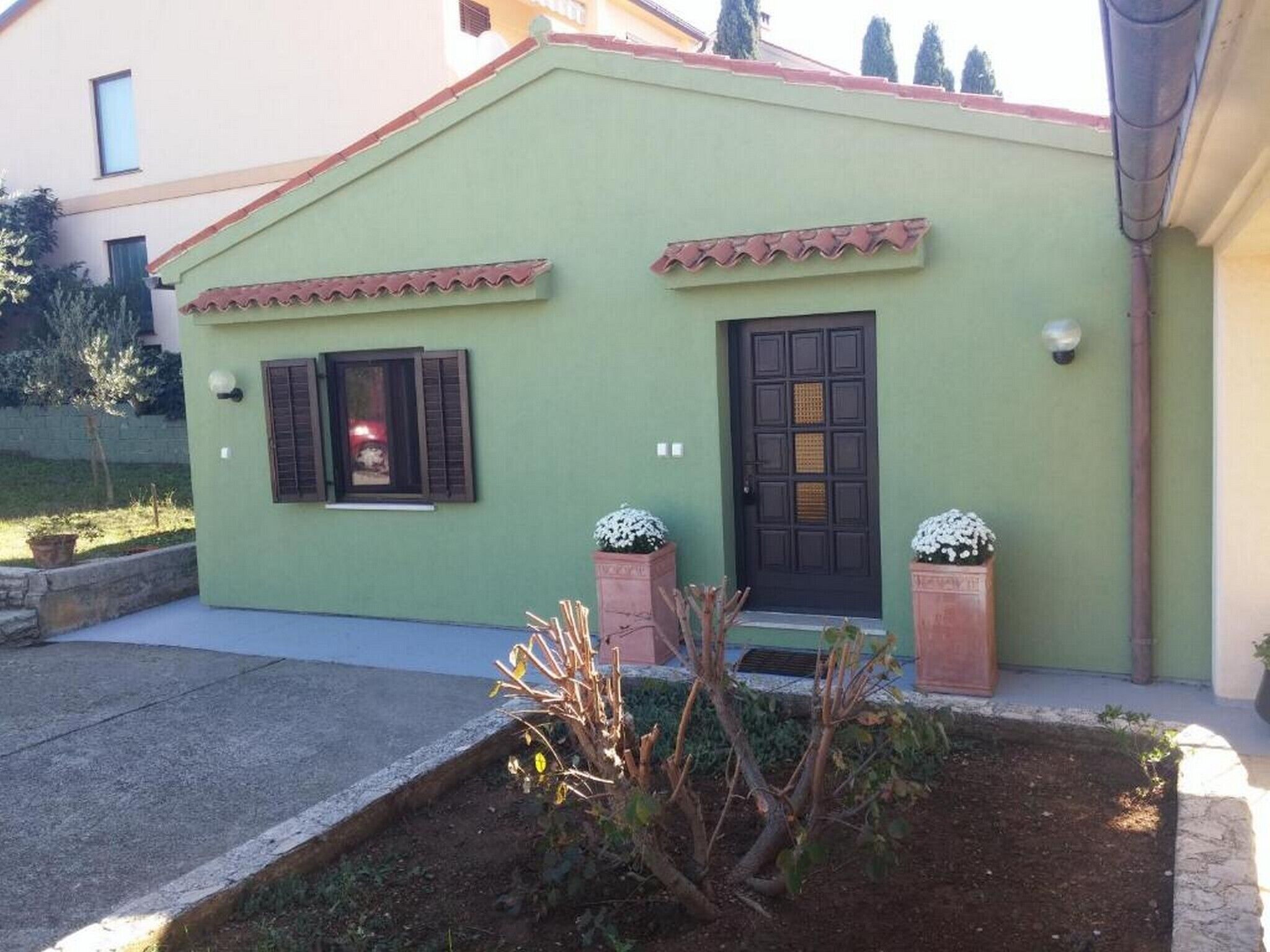 Una casa pequeña de un piso con fachada verde, una puerta delantera negra y dos ventanas con contraventanas. Hay plantas en macetas frente a la puerta.