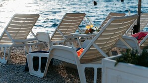 Strandstühle auf einer Kieselstreuung, dazwischen ein Tisch, mit Blick auf ein Gewässer.