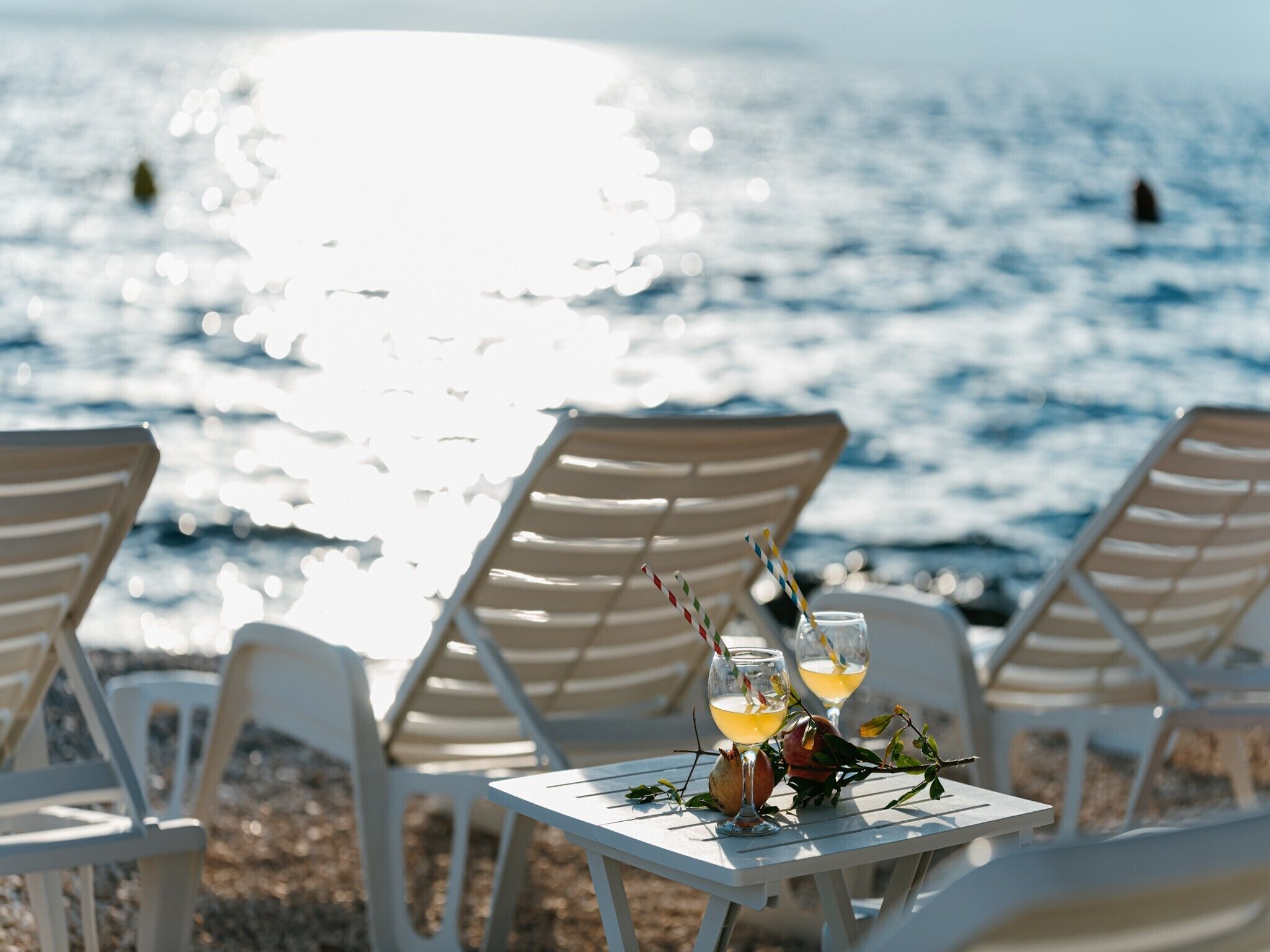 A table with two glasses of a yellow beverage, a pomegranate, and a branch with leaves, set against a backdrop of a beach with white chairs and a view of the sea.