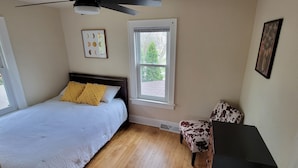 Una camera da letto con un letto, due finestre, una ventola a soffitto, una sedia e un quadro appeso al muro.