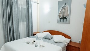 Ein ordentlich bezogenes Bett mit weißer Bettdecke und Kissen, ein kleiner Nachttisch mit Lampe sowie eine Wand mit einem Segelbootgemälde.