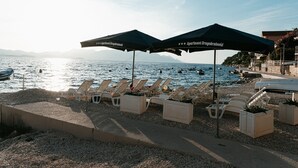 A beachside lounge with lounge chairs and umbrellas, overlooking a calm sea with boats and distant mountains. - Apartments Dragobratović - One Bedroom Apartment with Balcony and Sea View (Apartment 3) (Klek)