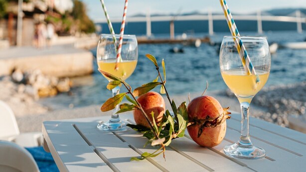 Two glasses of yellow beverage with striped straws on a white table by the sea, with a pomegranate and leaves nearby.