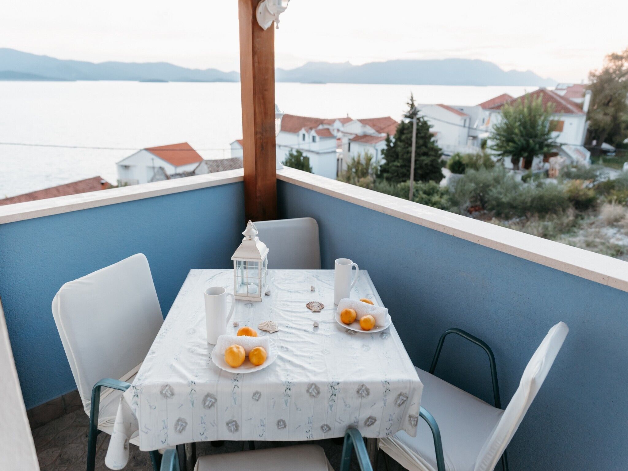 A balcony with a table set for two, featuring a white tablecloth, two chairs, a white lantern, a white mug, and a plate with oranges.