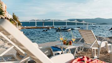 Vista de la playa con sillas de descanso, una mesa con bebidas y un puente al fondo.