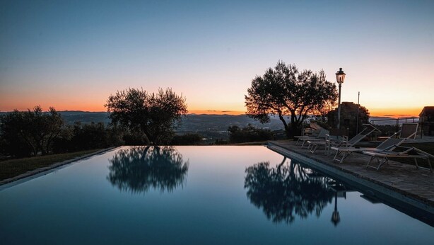 Una piscina a sfioro tranquilla che riflette alberi e un tramonto, con lettini e un faro.