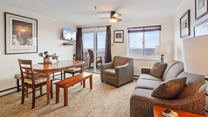 A living room with a wooden dining table, two chairs, a sofa, and a TV mounted on the wall.