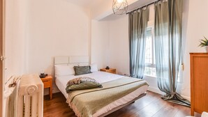 Et soveværelse med seng, sengeborde, en radiator, et vindue med gardiner og en hængende lysarmatur.
