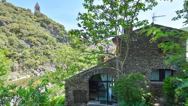 A stone building with a balcony overlooking a river, surrounded by lush greenery and a hill with a tower in the background. - La Mazette, les pieds dans l'eau (Olargues)