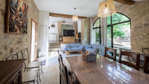 A spacious dining area with a wooden table and chairs, a sofa, and a large window offering a view of the outdoors. - La Mazette, les pieds dans l'eau (Olargues)