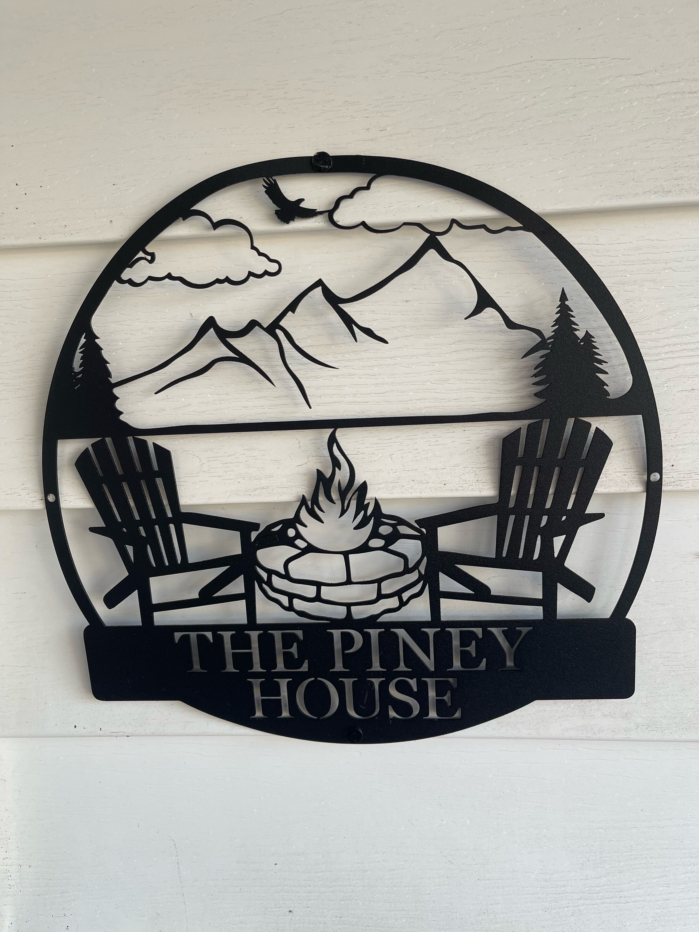 Un panneau circulaire représentant un paysage de montagne, deux chaises Adirondack, un feu de camp et l’inscription « The Piney House ».