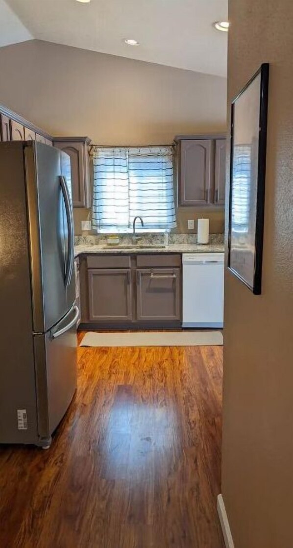 A compact kitchen with wooden flooring, a refrigerator, a dishwasher, and a window with blinds.