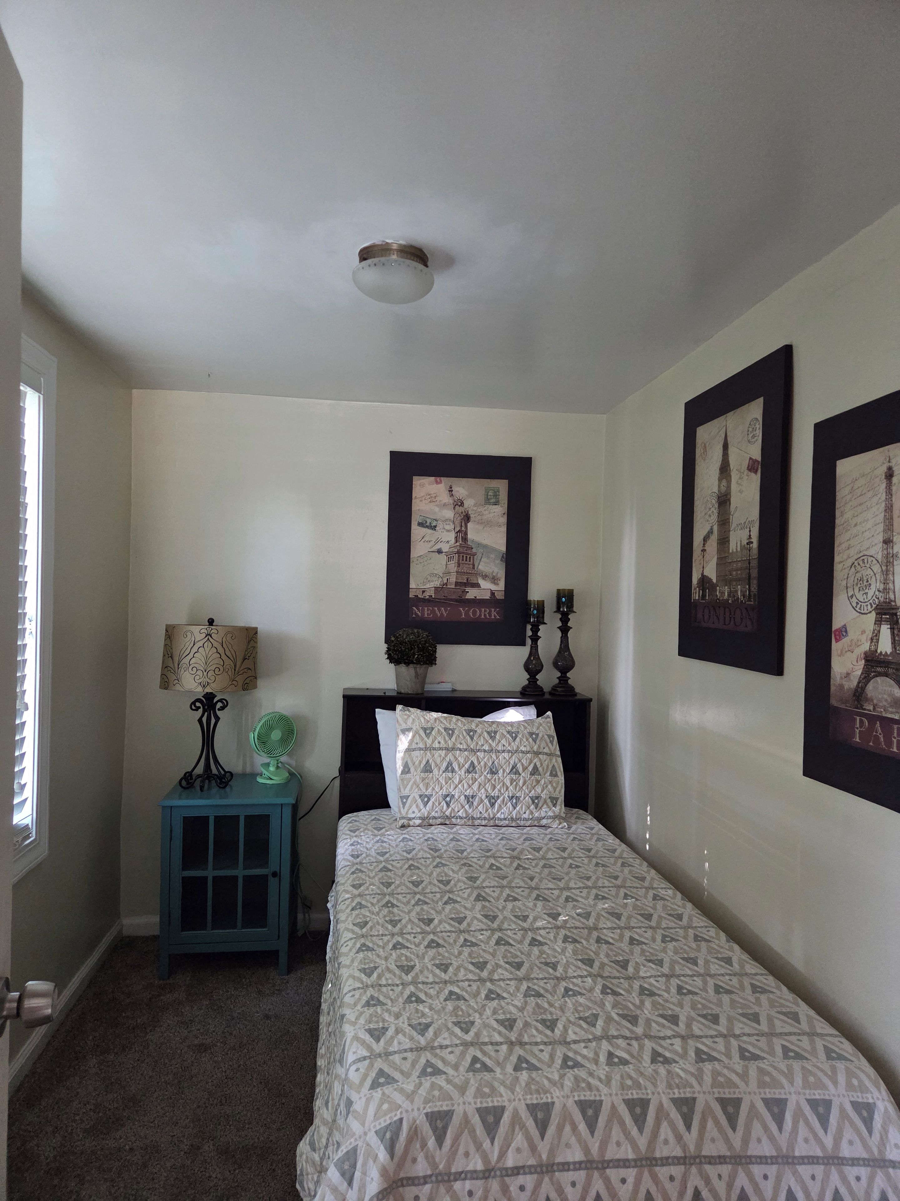 A bedroom with a bed, a nightstand with a lamp, a small table with a fan, and two framed pictures on the wall.