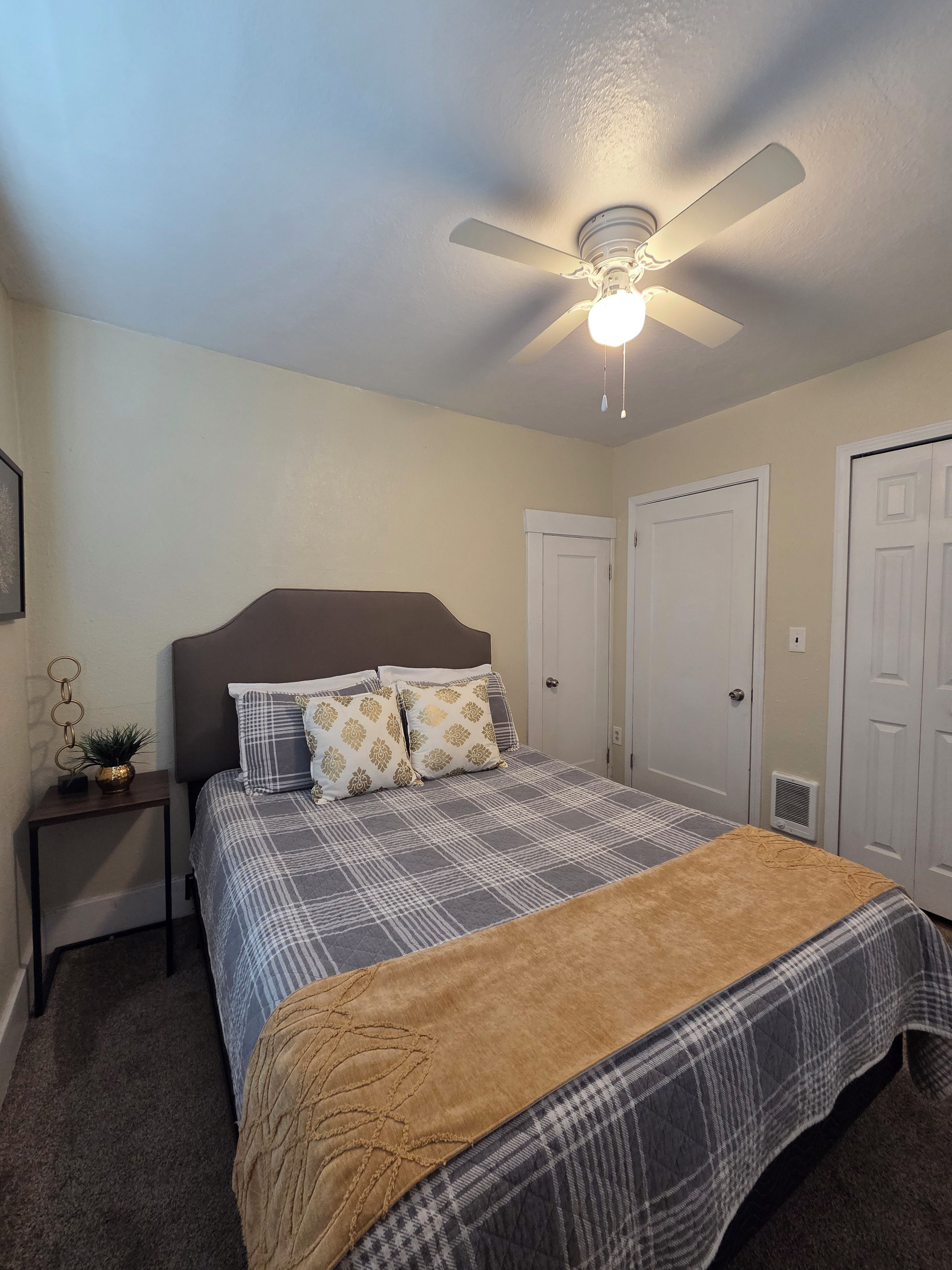 Quarto com cama, ventoinha no teto, mesa de cabeceira e porta.