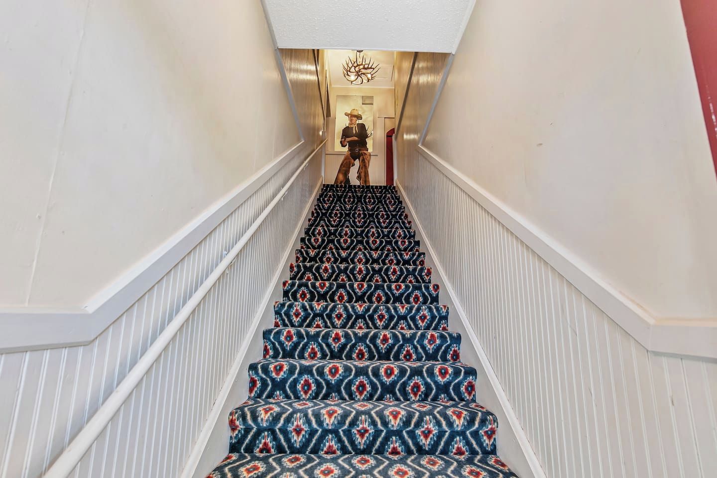 A staircase with patterned carpet and white railings leading to a room with a chandelier.