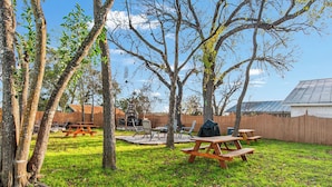 An outdoor patio area with picnic tables and a barbecue grill, surrounded by trees and a wooden fence. - Carriage Utopia 4 | Pet-Friendly Stay in Bandera (Bandera)
