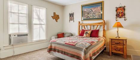A bedroom with a large bed, a wooden headboard, a nightstand with a lamp, a wall painting, and a decorative wall hanging.
