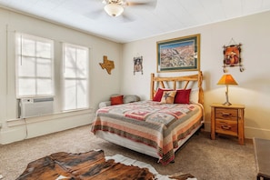 A bedroom with a large bed, a wooden headboard, a nightstand with a lamp, a wall painting, and a decorative wall hanging.