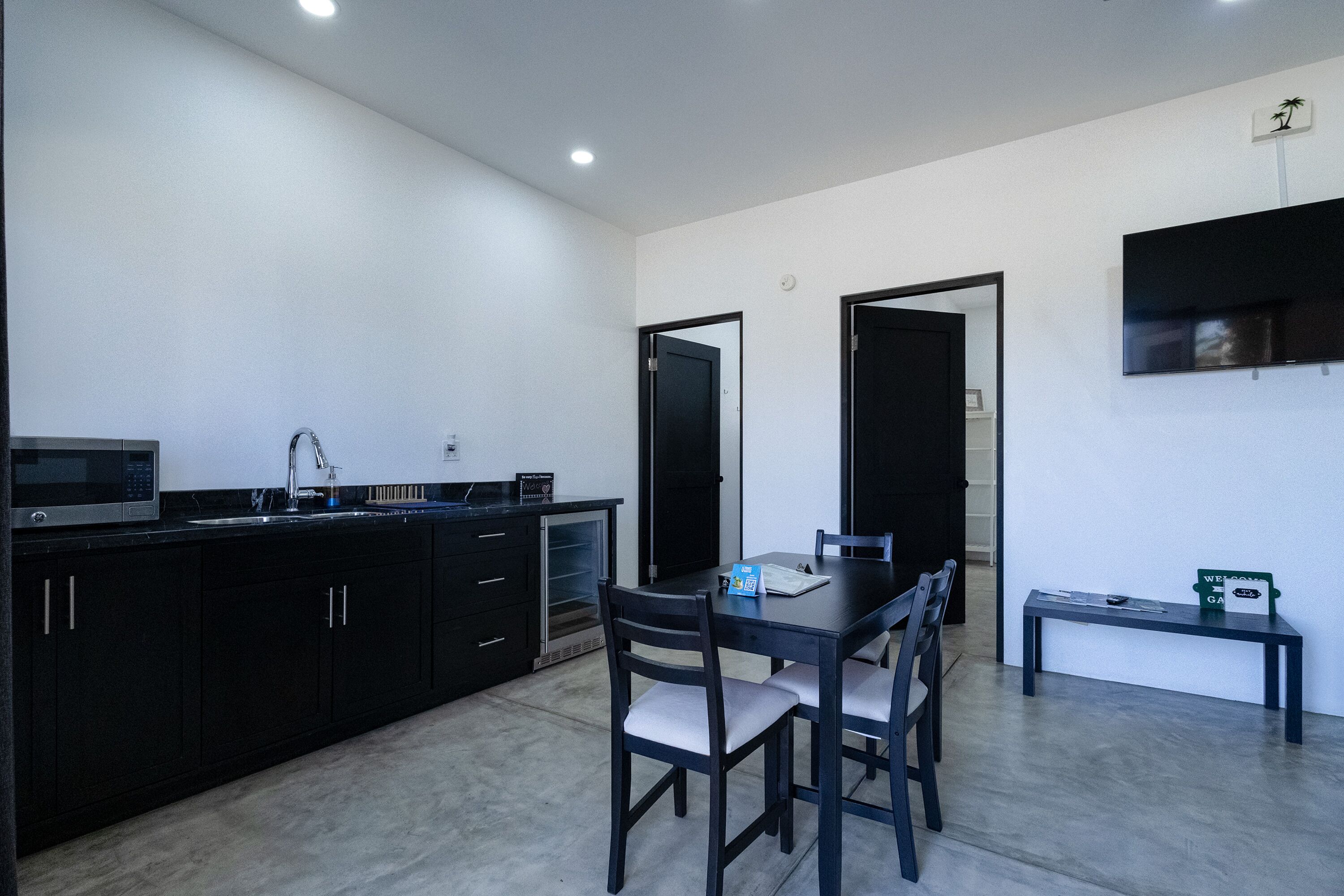 A modern kitchen with black cabinets, a dining table with four chairs, a microwave, and a TV mounted on the wall.