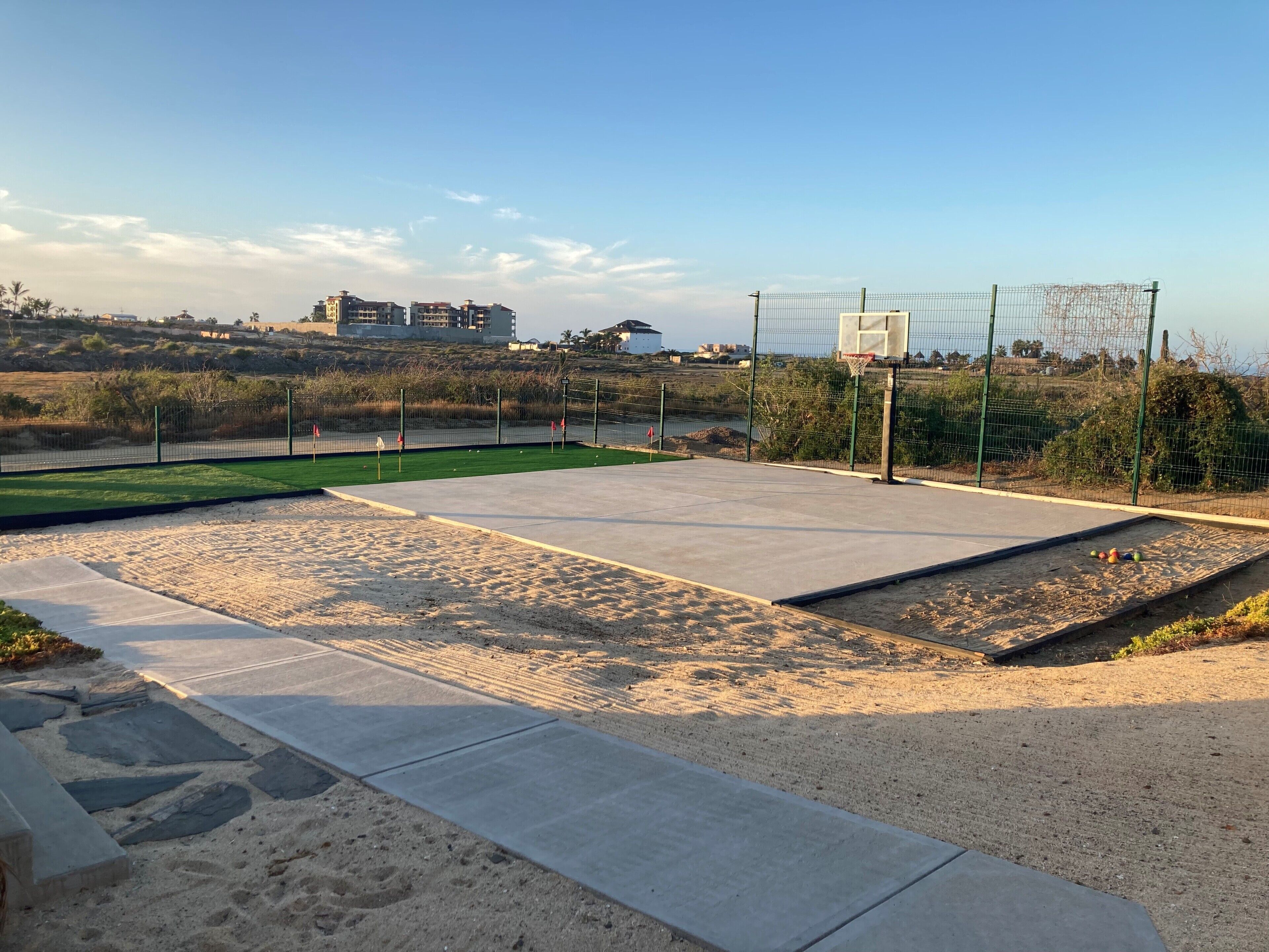 Una cancha de baloncesto al aire libre con aro, rodeada por una cerca y una zona de arena.