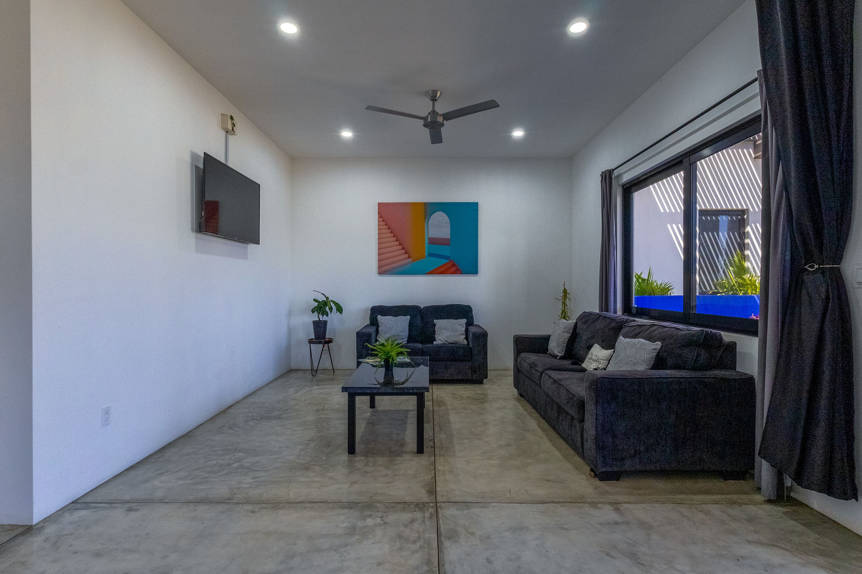 A living room with a ceiling fan, a flat-screen TV, a colorful abstract painting, two sofas, a coffee table, and a large window with curtains.