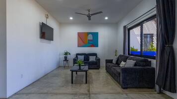 A living room with a ceiling fan, a flat-screen TV, a colorful abstract painting, two sofas, a coffee table, and a large window with curtains.