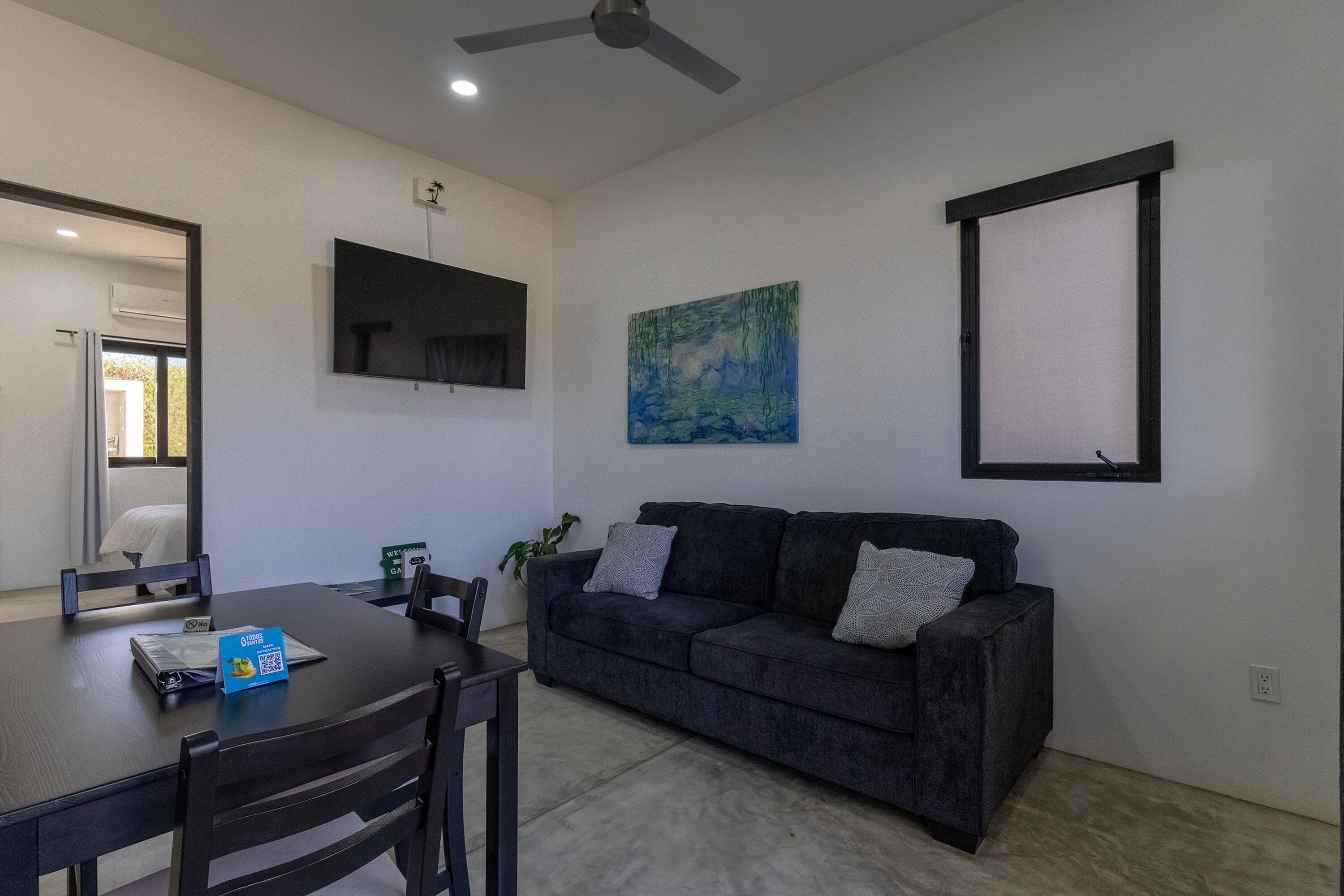 A modern living room with a dark grey sofa, a flat-screen TV mounted on the wall, a painting, and a dining area with a table and chairs.