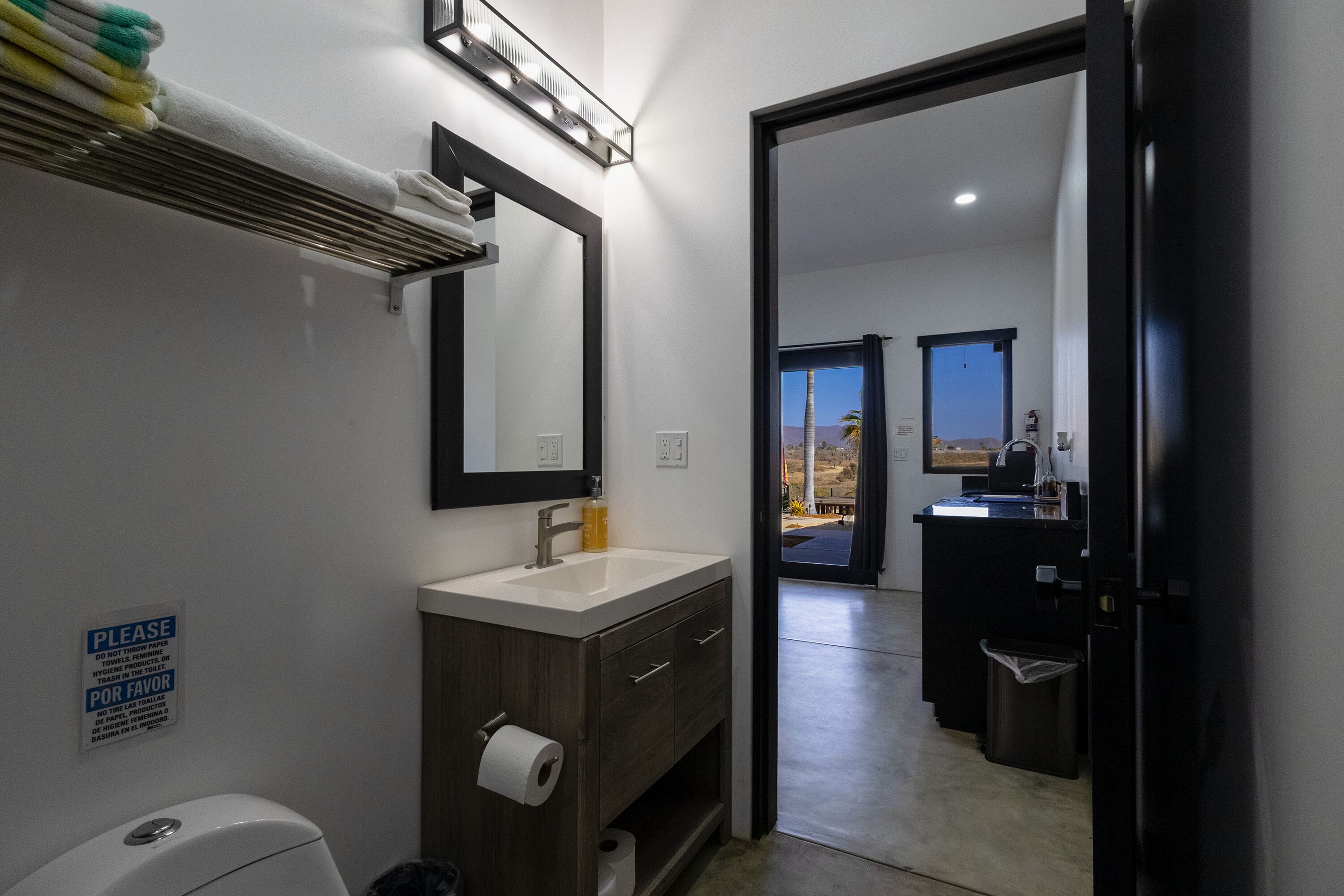 A bathroom with a sink, mirror, towel rack, and a sign instructing not to throw trash in the toilet.