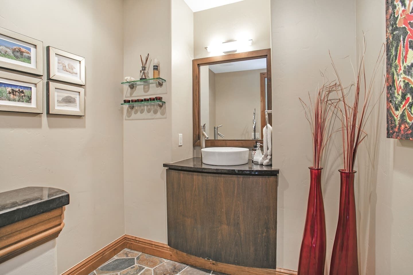 Un baño con tocador de madera, un lavabo blanco y un espejo. Hay cuadros enmarcados en la pared, una repisa de vidrio con artículos de tocador y dos jarrones rojos.