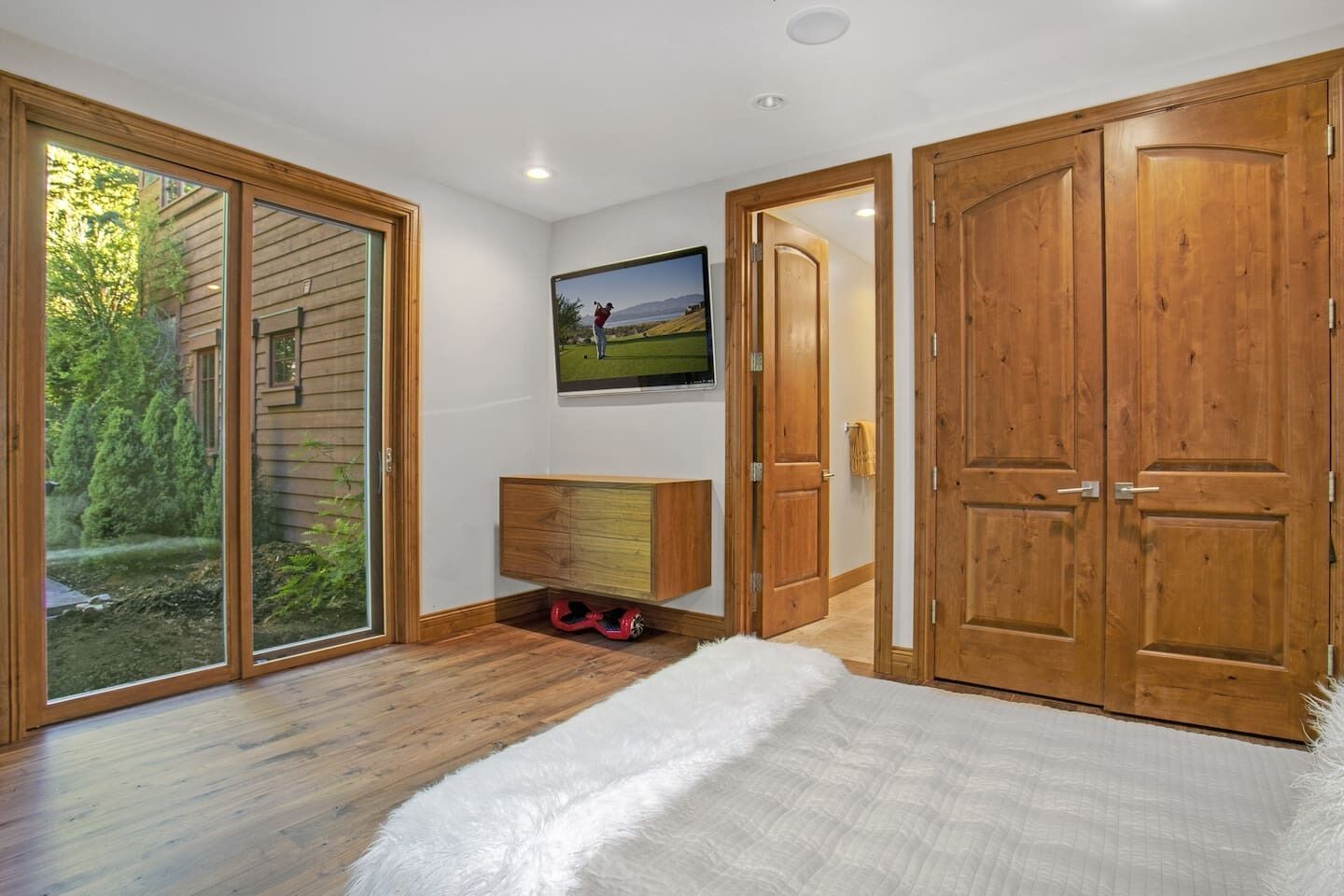 A bedroom with a large sliding glass door, a wooden wardrobe, a TV mounted on the wall, and a fluffy white bedspread.