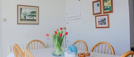 A dining area with a table set for two, surrounded by wooden chairs. A vase with red tulips and a blue teapot are on the table.