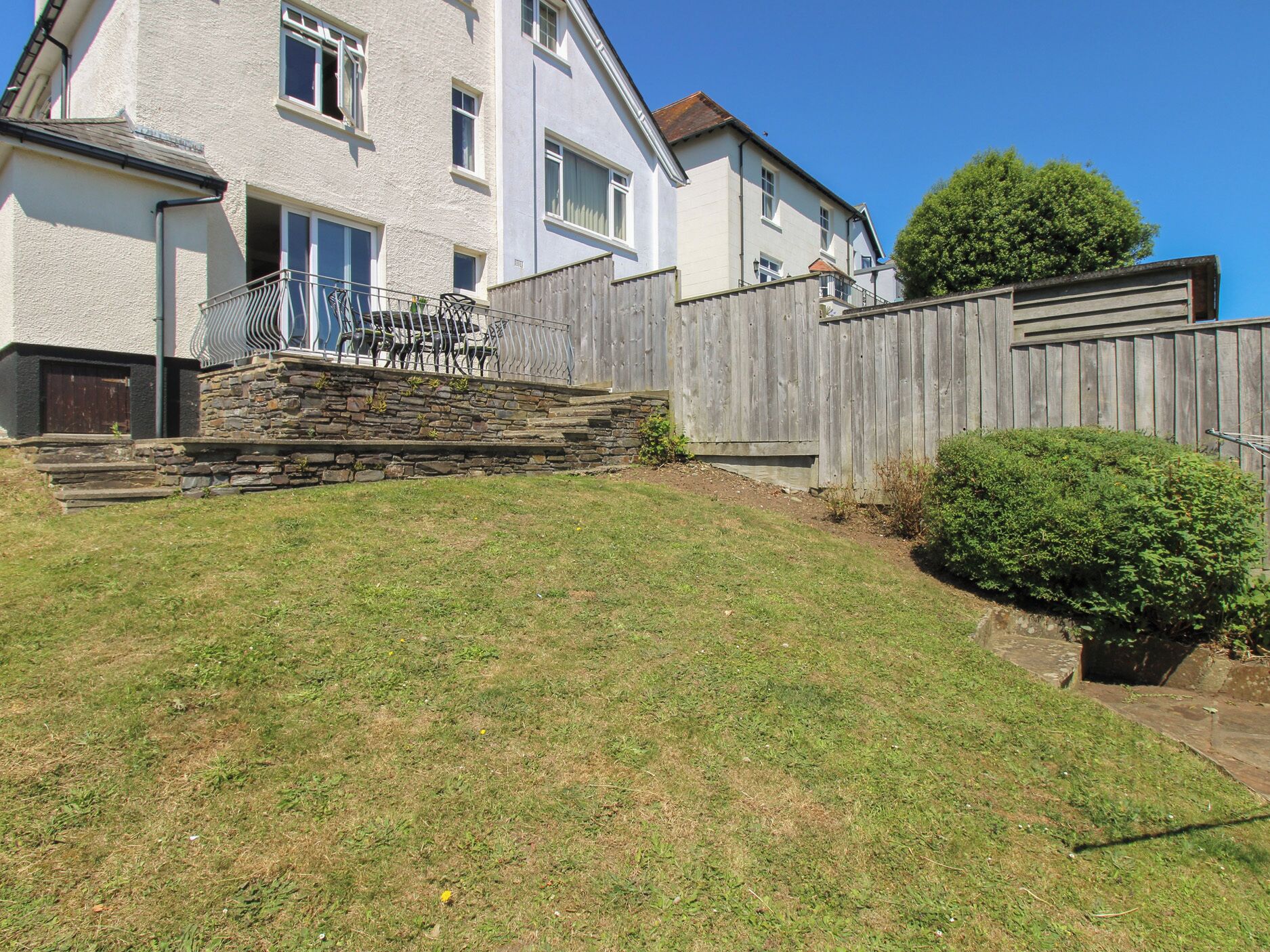 Une maison à deux étages avec un escalier en pierre menant à un balcon, une clôture en bois et une pelouse bien entretenue.