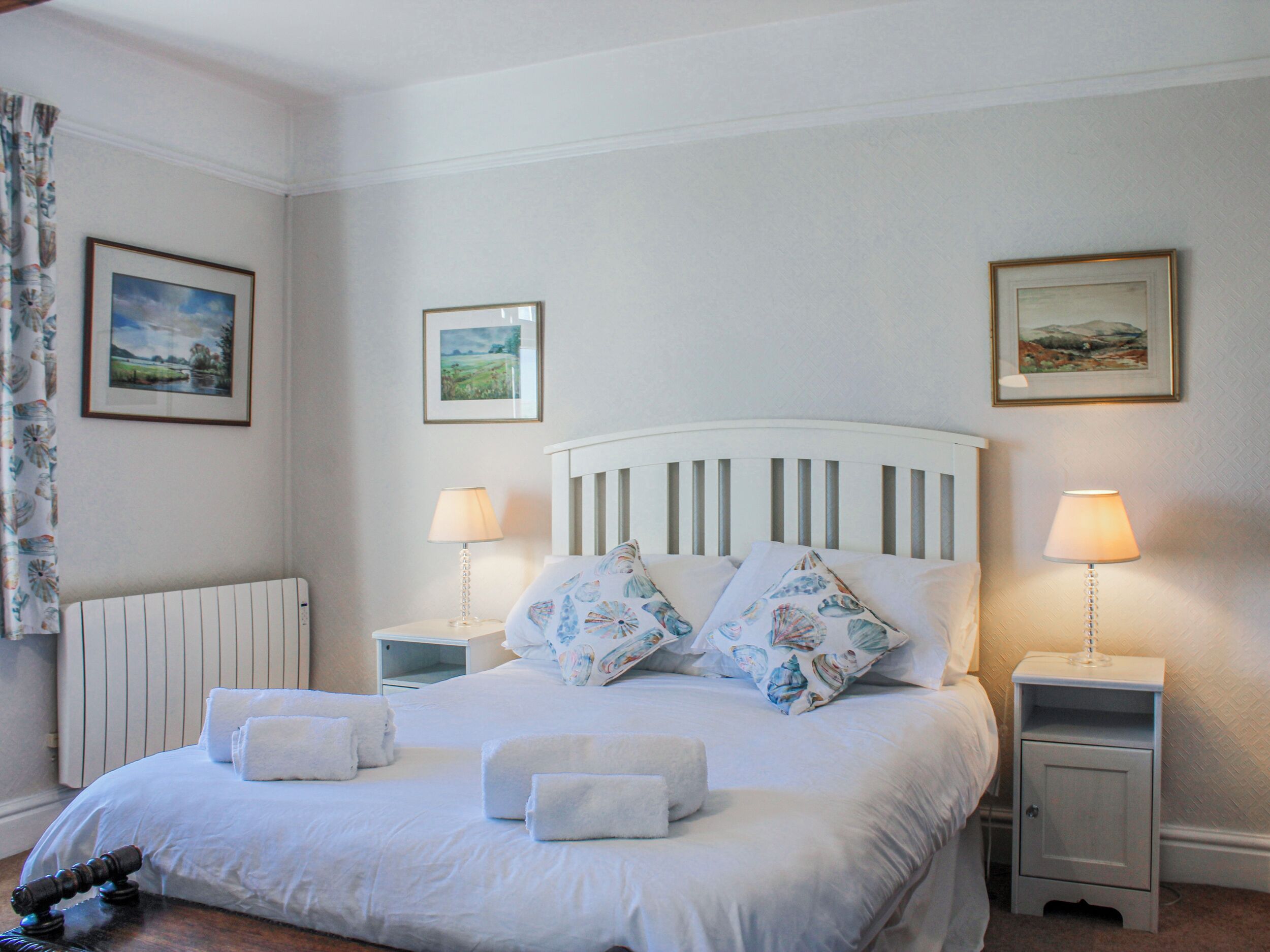 A neatly made bed with white linens and floral pillows, two bedside tables with lamps, and framed landscape paintings on the walls.