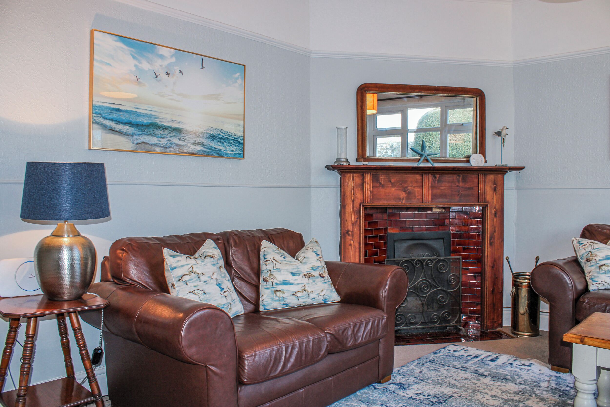 A living room with a brown leather sofa, a blue rug, a fireplace, and a painting of a seascape.