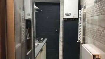 Un couloir étroit, avec un coin cuisine sur la gauche, une porte noire au fond et un radiateur sur la droite.