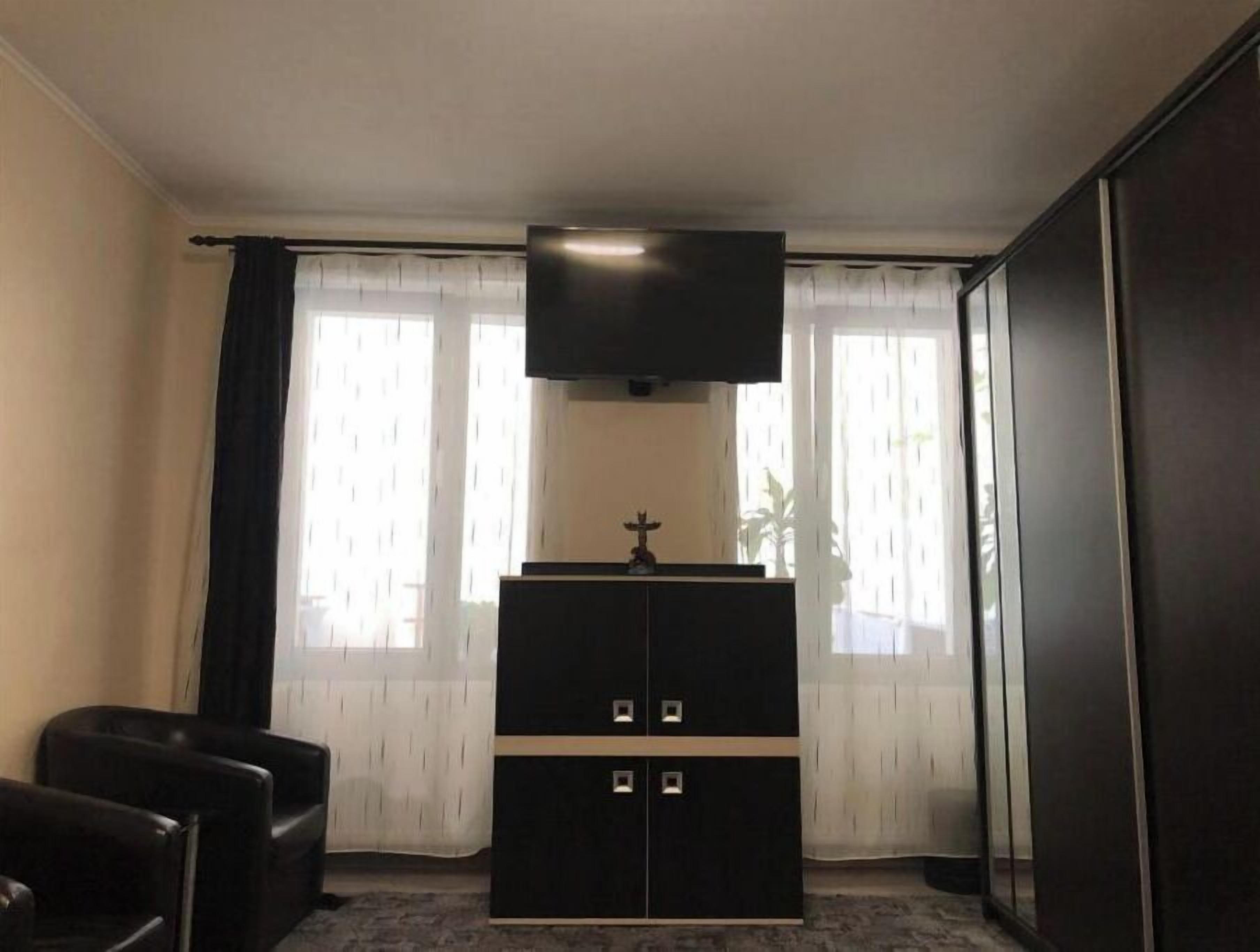 Une pièce avec une armoire en bois sombre, un téléviseur à écran plat fixé au-dessus, et une petite statue posée sur le dessus. On y trouve deux fauteuils en cuir noir et une grande fenêtre ornée de rideaux légers.