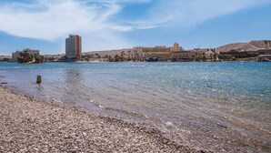 Eine Küstenlandschaft mit Kiesstrand, klarem Wasser und einem großen Hotel im Hintergrund.
