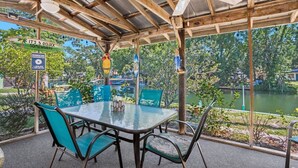 Un patio exterior con una mesa y sillas de vidrio, rodeado por una pérgola de madera con ventilador de techo.