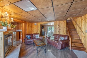 A cozy cabin interior with wooden walls, leather sofas, a fireplace, and a staircase.
