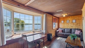 A wooden interior with a dining table, chairs, a sofa, and a view of a lake through large windows. - Rustic Riverside Charm and Exclusive Fishing Access! (Richland)