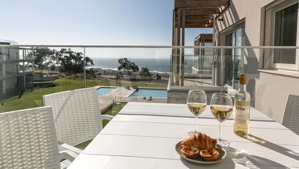Un balcon avec une table dressée pour deux personnes, agrémentée d’un croissant et de verres à vin, offrant une vue sur une piscine et l’océan.