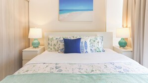 Een net opgemaakt bed met een bloemenbedekking, twee bijpassende lampen en een schilderij met een strandthema aan de muur.